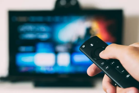Hand holding a TV remote in a room, aimed at a large screen showing a blurred grid of streaming thumbnails with blue wall glow.